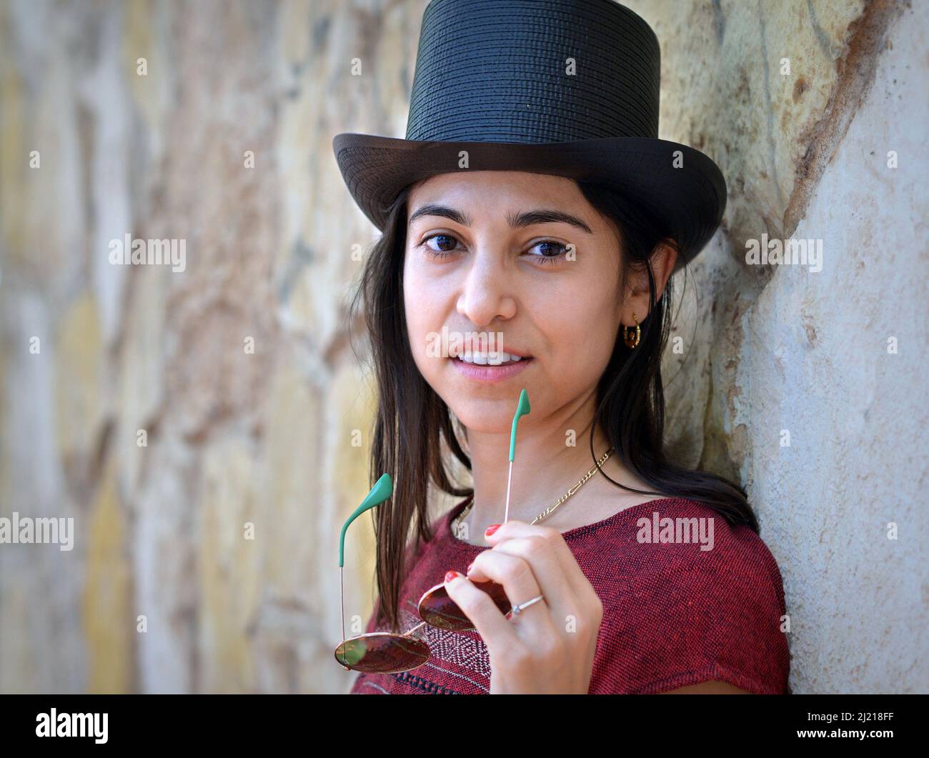 Die junge, schöne, charmante Latina-Frau trägt einen schwarzen Zylinderhut, hält eine Sonnenbrille und lächelt den Betrachter vor einem Hintergrund aus einer Steinwand an. Stockfoto