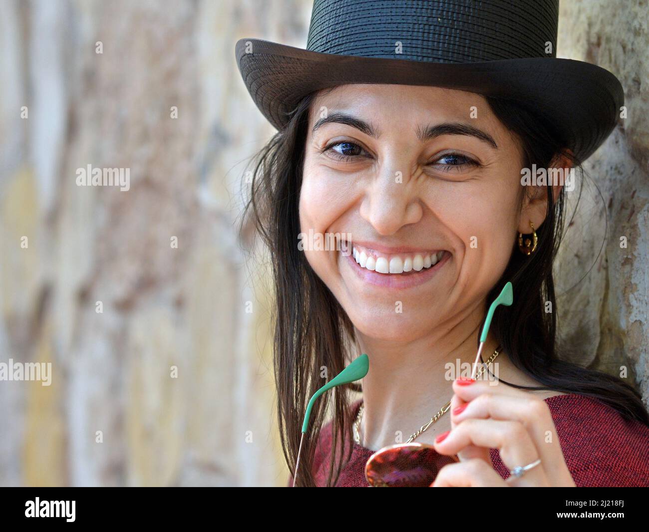 Die junge, schöne, charmante Latina-Frau trägt einen schwarzen Zylinderhut, hält eine Sonnenbrille und lächelt den Betrachter vor einem Hintergrund aus einer Steinwand an. Stockfoto