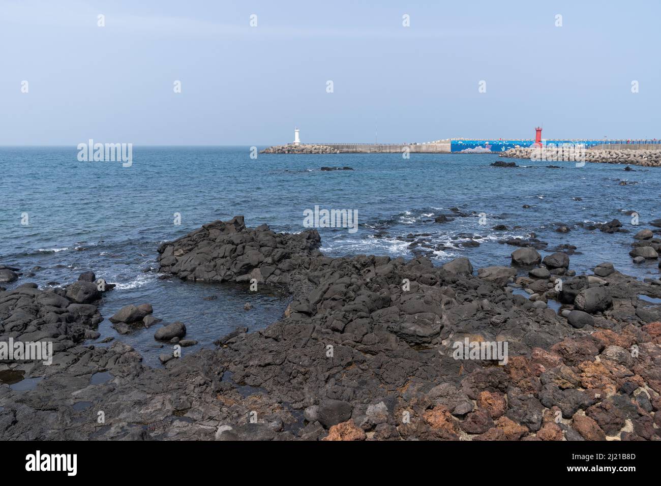 Schwarzes vulkanisches Gestein im Hafen von Mosulpo auf der Insel Jeju, Südkorea. Zwei Leuchttürme und ein buntes Wandbild sind auf den Betonbrechern dahinter zu sehen. Stockfoto