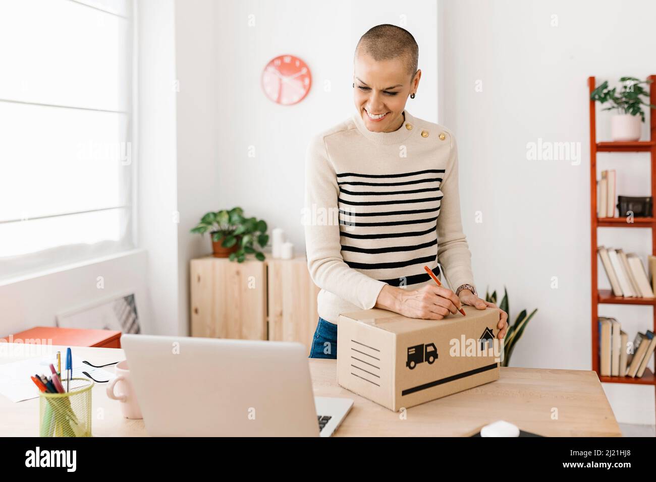 Junge Frau, die im Heimbüro mit einem Laptop arbeitet, während sie auf einem Karton schreibt Stockfoto