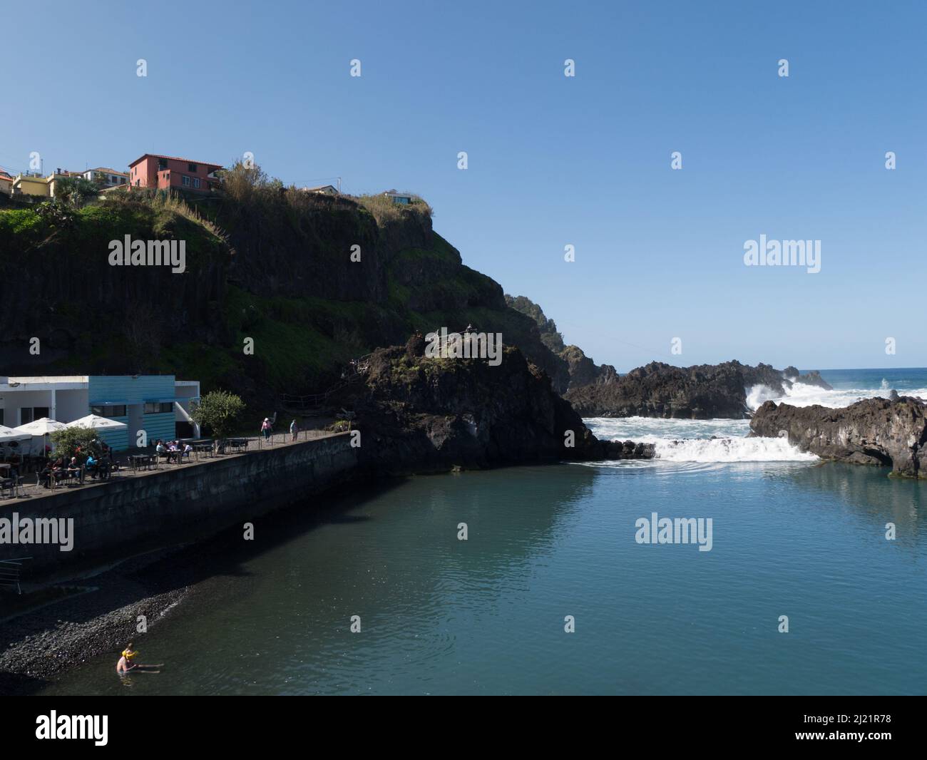 Besucher sitzen in Sonnenschein im Café an der Küste von Seixal Madeira Portugal EU mit Wellen des Atlantischen Ozeans, die über vulkanischem Gestein krachen Stockfoto