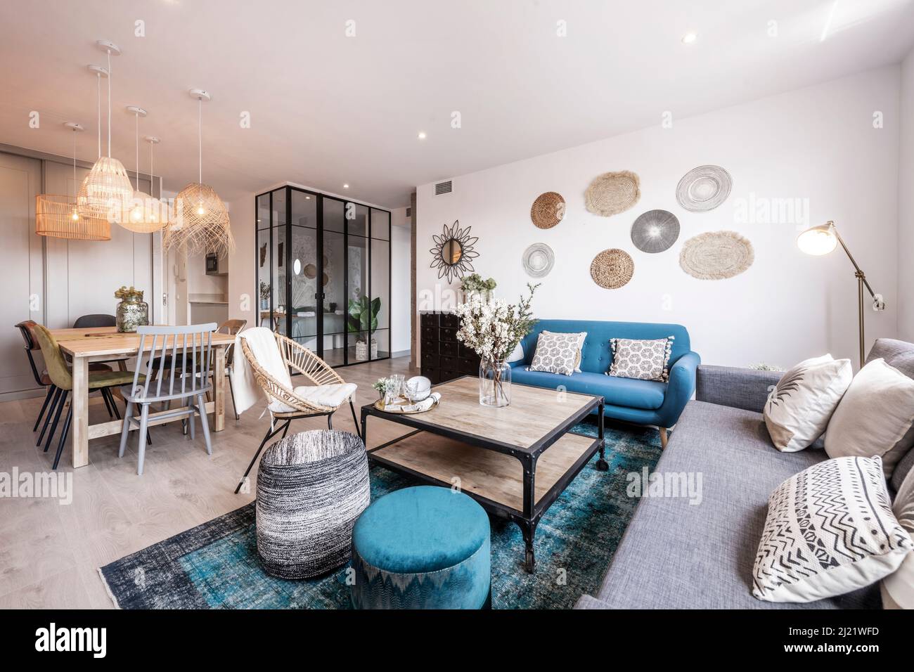 Wohnzimmer mit Esstisch aus Holz, gepolsterten Stühlen aus Holz und Metall und Korblampen, hellem Holzboden und blauem Sofa mit Teppich und schwarzem Metall Stockfoto