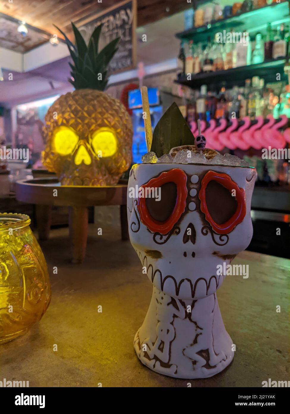 Ein Tiki-Cocktail-Getränk in einem Keramikbecher mit Zuckerschädel auf einer Bar. Hinter dem Getränk ist ein Ananas-Totenkopf-Licht. Stockfoto