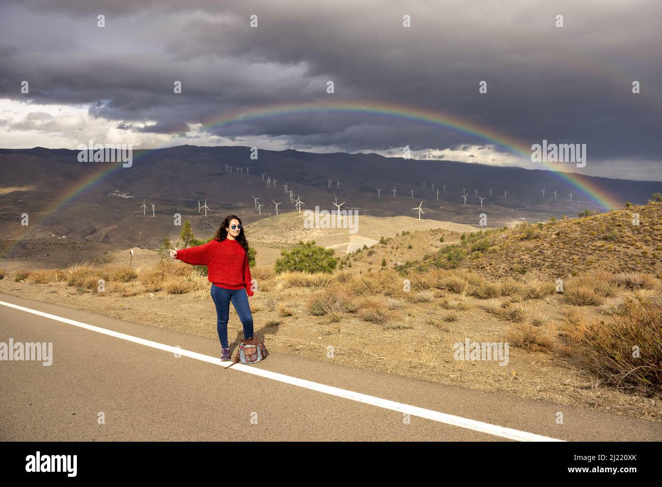 Frau in ihrem 25s Anhalter an einem Tag mit Sonne, Wolken und Regenbögen im Hintergrund. Stockfoto