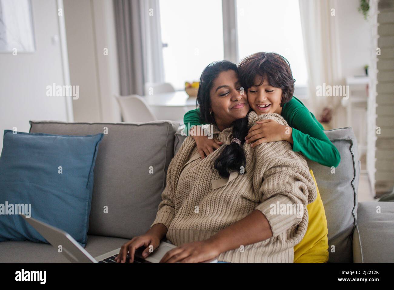 Die indische Mutter benutzt einen Laptop und ihr kleiner Sohn umarmt sich zu Hause auf dem Sofa. Stockfoto