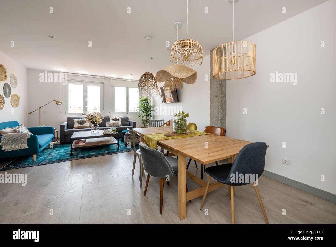 Wohnzimmer mit einem Esstisch aus Holz, gepolsterten Stühlen aus Holz und Metall und Korblampen, hellen Holzböden und einem blauen Sofa mit Teppich Stockfoto