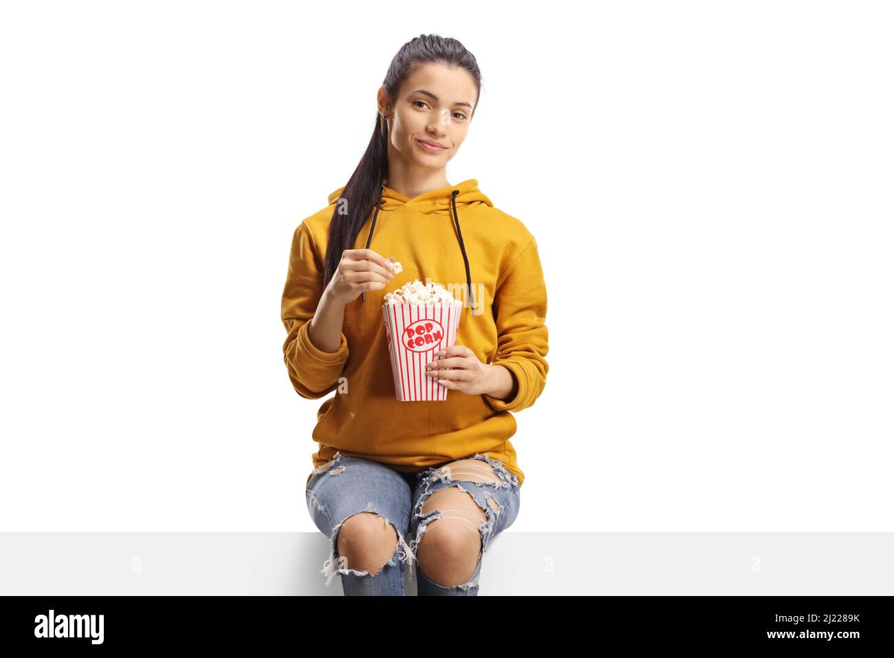 Junge Frau, die auf einer Tafel sitzt und auf weißem Hintergrund eine Schachtel Popcorn isoliert hat Stockfoto