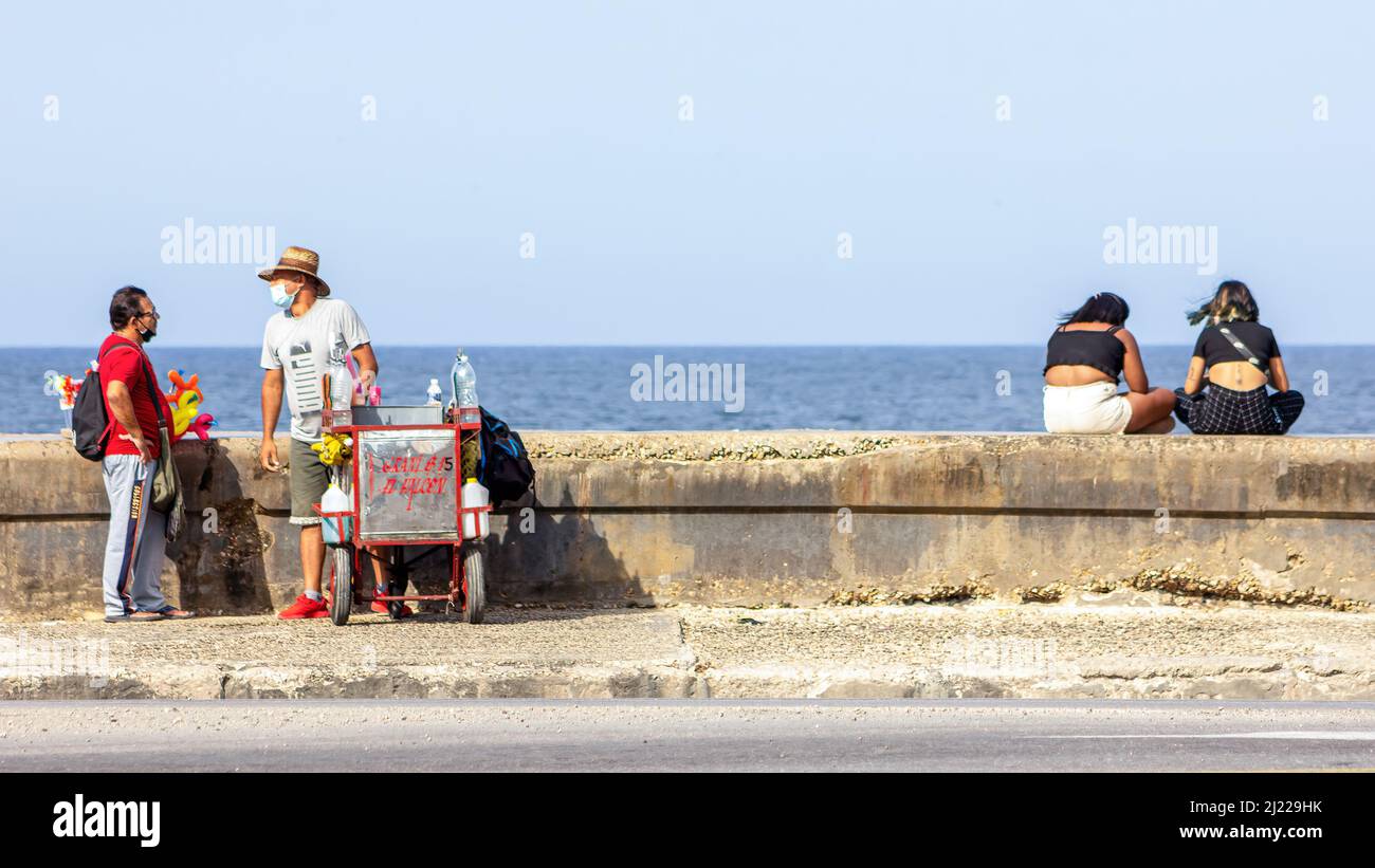 Ein selbstständiger Kubaner mit Gesichtsmaske verkauft in El Malecon aromatisiertes Eis. Er spricht mit einem anderen Mann, während zwei junge Frauen im se sitzen Stockfoto