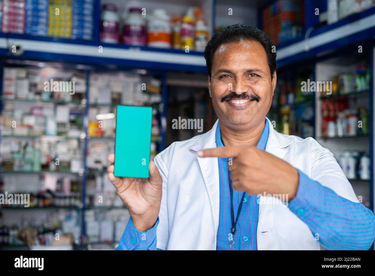 Lächelnder Apotheker in Uniform zeigt grünes Handy, indem er mit dem Finger auf die Apotheke zeigt - Konzept der Online-Buchung, Werbung Stockfoto