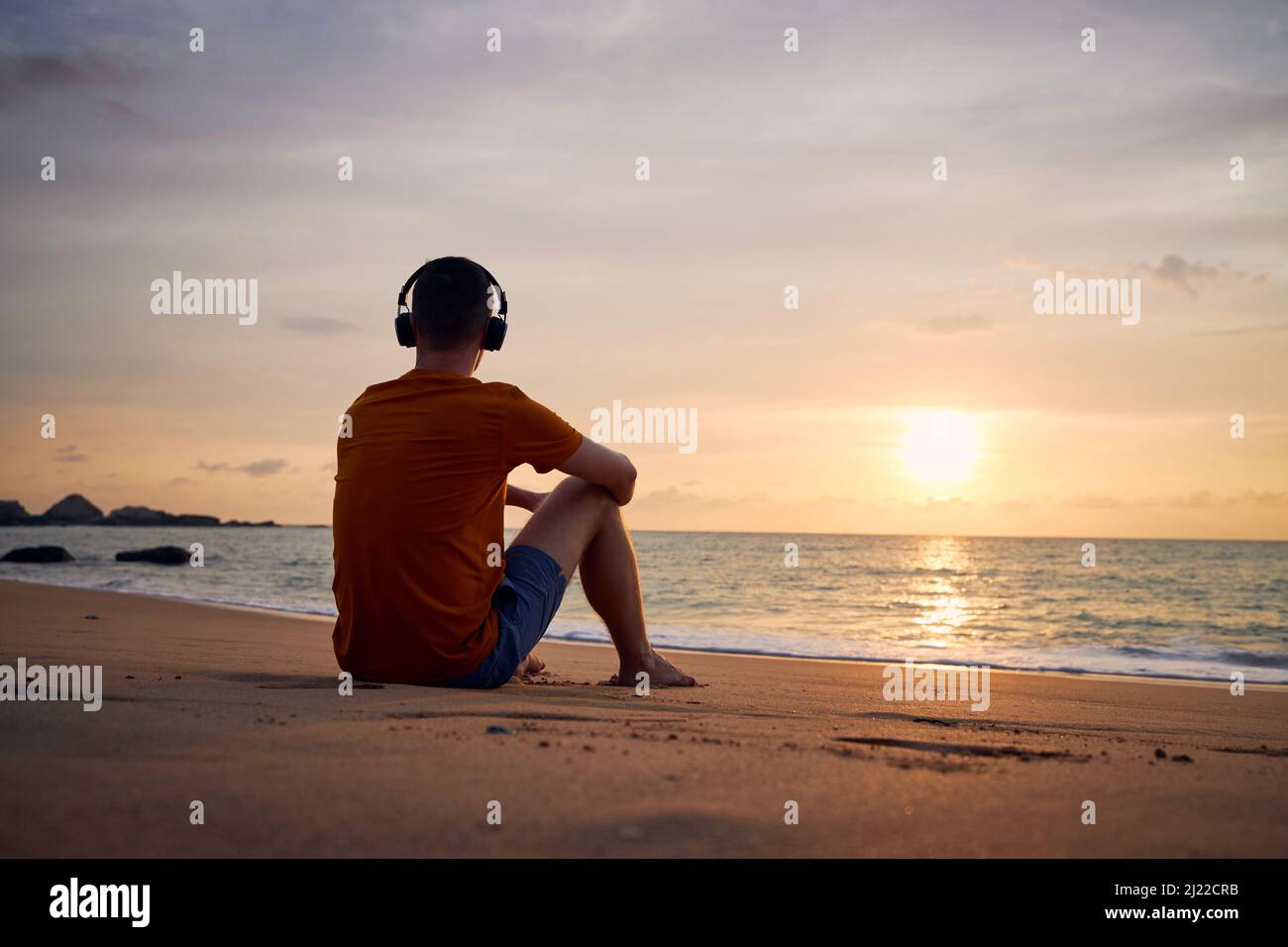 Rückansicht des Mannes mit Kopfhörern am Strand. Gelassenheit, Kontemplation und Musikhören bei schönem Sonnenuntergang. Stockfoto