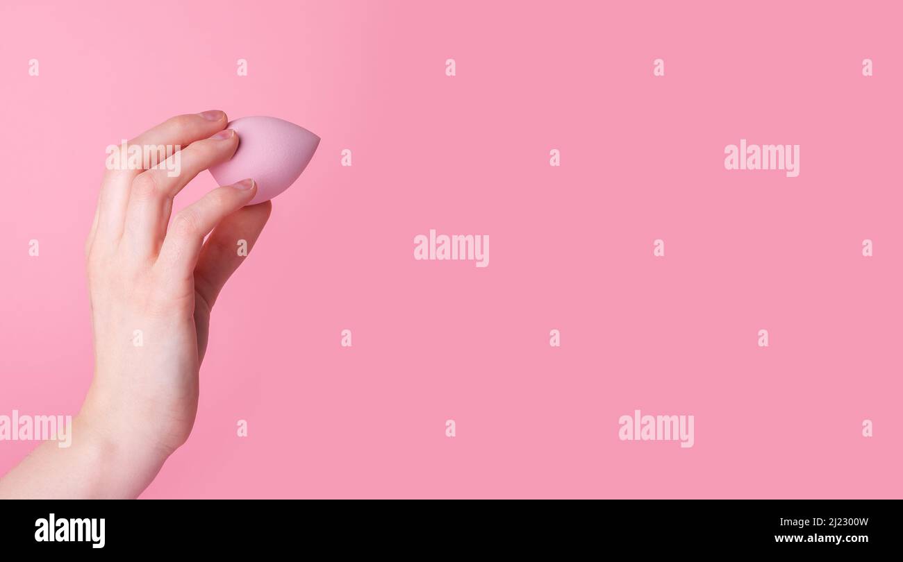 Banner mit Hand hält Make-up Schwamm oder Mixer auf rosa Hintergrund. Schönheitskonzept. Auftragen von Fundament, Concealer, Balsam. Gesichtskonturierung. Speicherplatz kopieren. Hochwertige Fotos Stockfoto