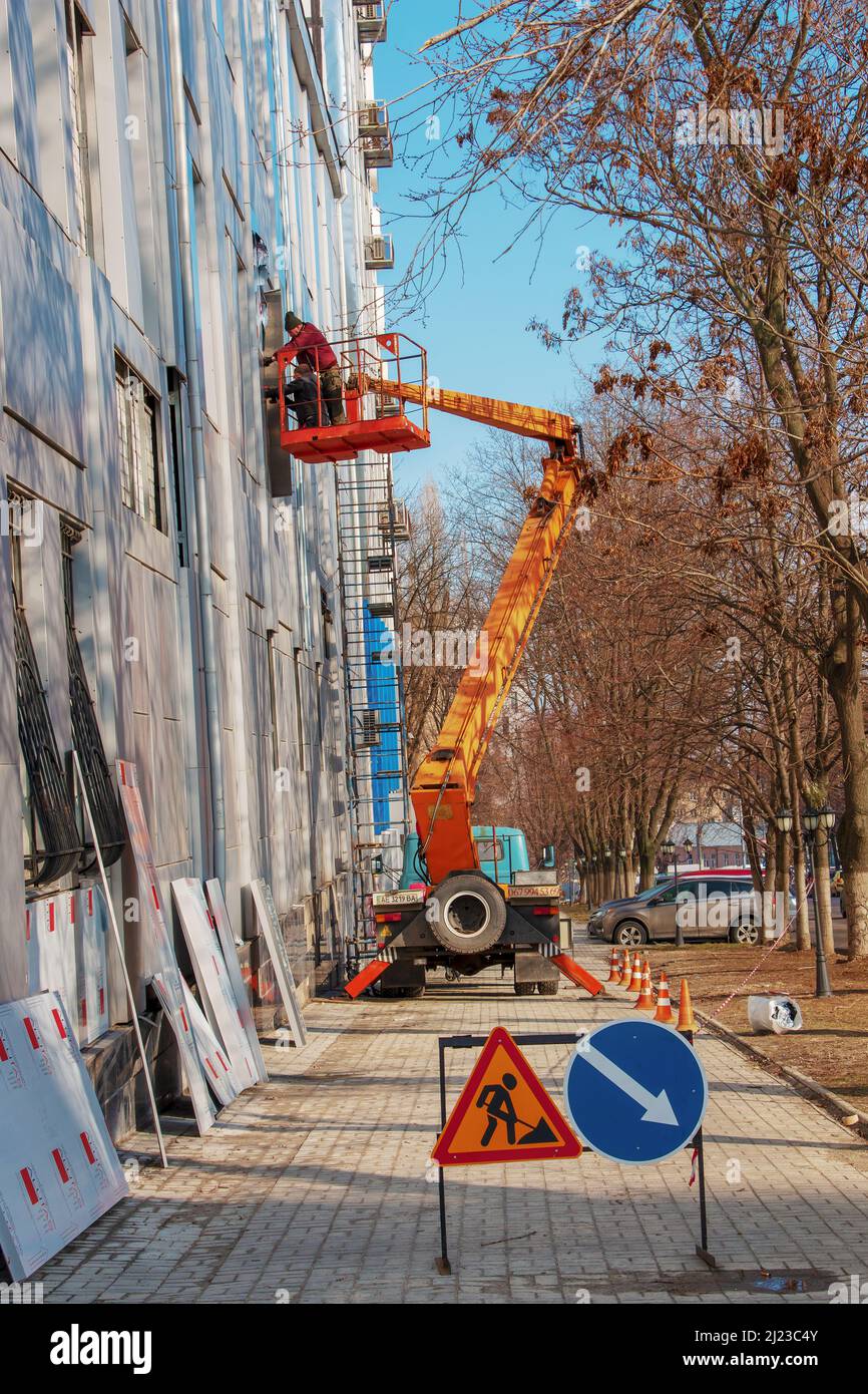 Dnepropetrovsk, Ukraine - 02.09.2022: Baumeister in einem Krankorb repariert die Beschichtung einer Metallstruktur an der Wand der Fassade eines Gebäudes. Stockfoto