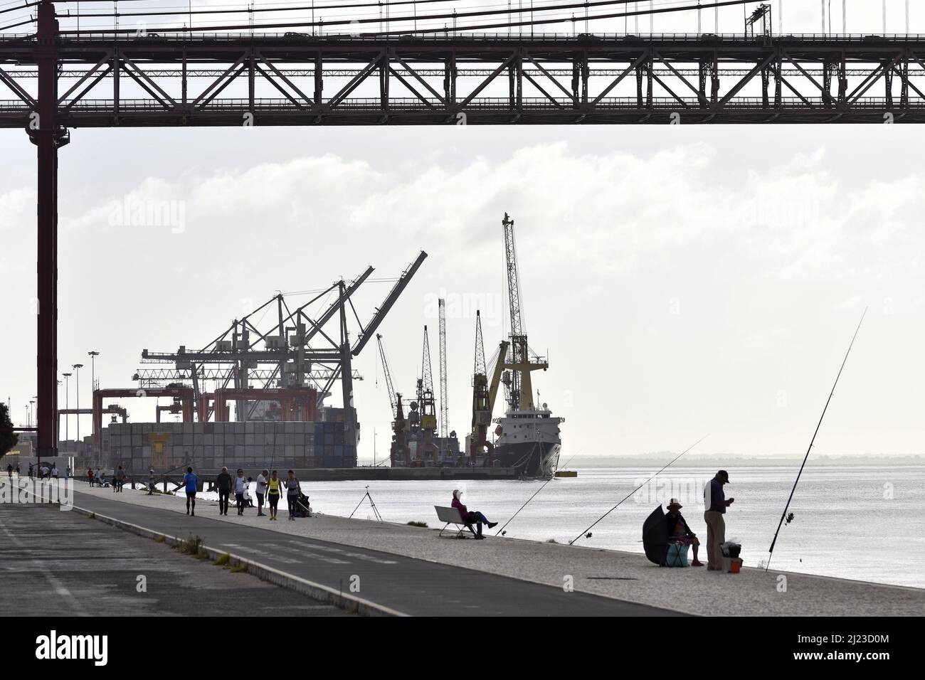 Menschen an der Promenade und Fischer am Fluss Tejo in der Nähe der Brücke 25 de Abril mit Industriehafen im Hintergrund, Lissabon Portugal. Stockfoto