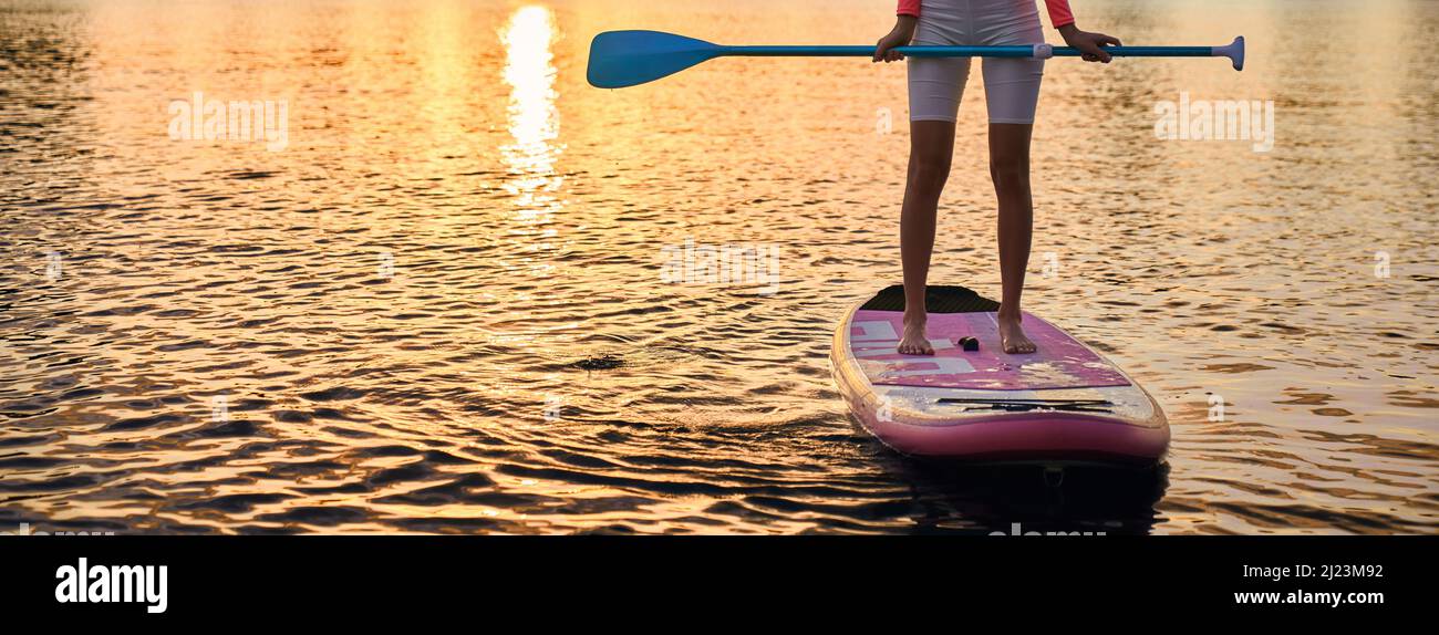 Nahaufnahme einer jungen Frau mit schlanken Beinen, die auf dem Brett mit langem Paddel in den Händen steht. Aktive Frau in Sportbekleidung, die abends Zeit für Wassersport verbringt. Stockfoto