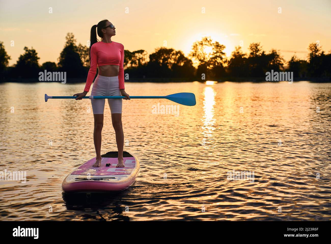 Attraktive Frau mit schlankem Körper schwimmt auf supboard während der Abendzeit. Kaukasische Brünette mit Paddel in den Händen Training an der frischen Luft mit erstaunlichen Sonnenuntergang auf dem Hintergrund. Stockfoto