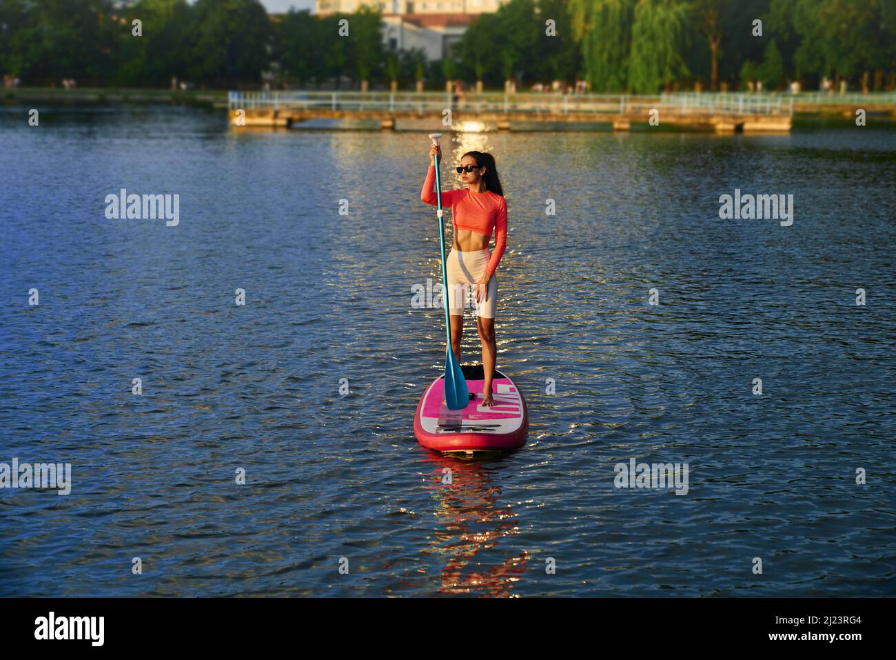 Athletische kaukasische Frau, die während des herrlichen Sonnenuntergangs im Sup-Surfen mit Paddel praktiziert. Junge Hündin schwimmend auf dem Stadtersee während der Abendzeit aktiv. Stockfoto