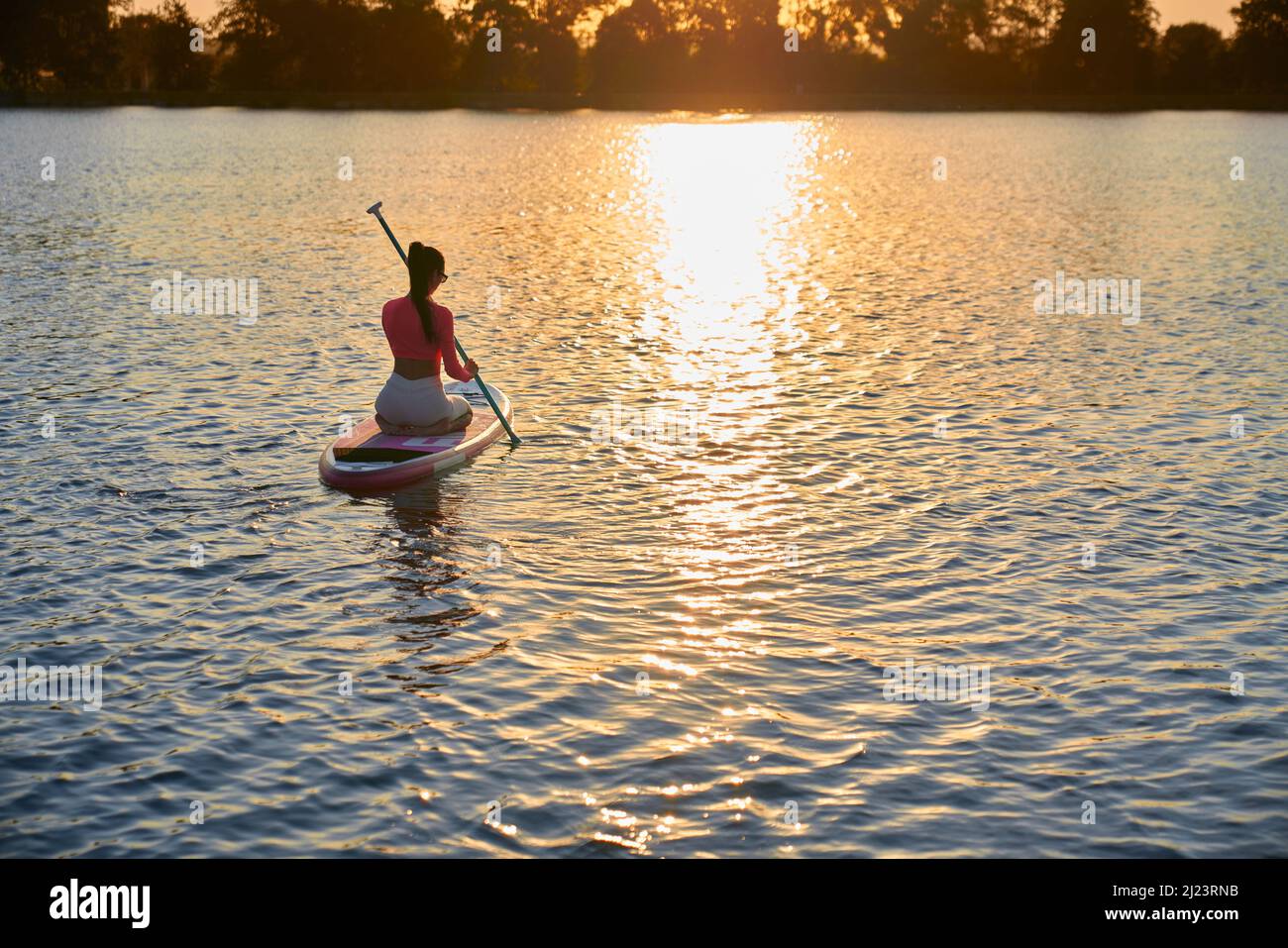 Aktive kaukasische Frau in bunten Sportkleidung beim Schwimmen mit Paddel auf dem Brett während der Abendzeit. Toller Sommeruntergang über dem See. Wassersport-Konzept. Stockfoto