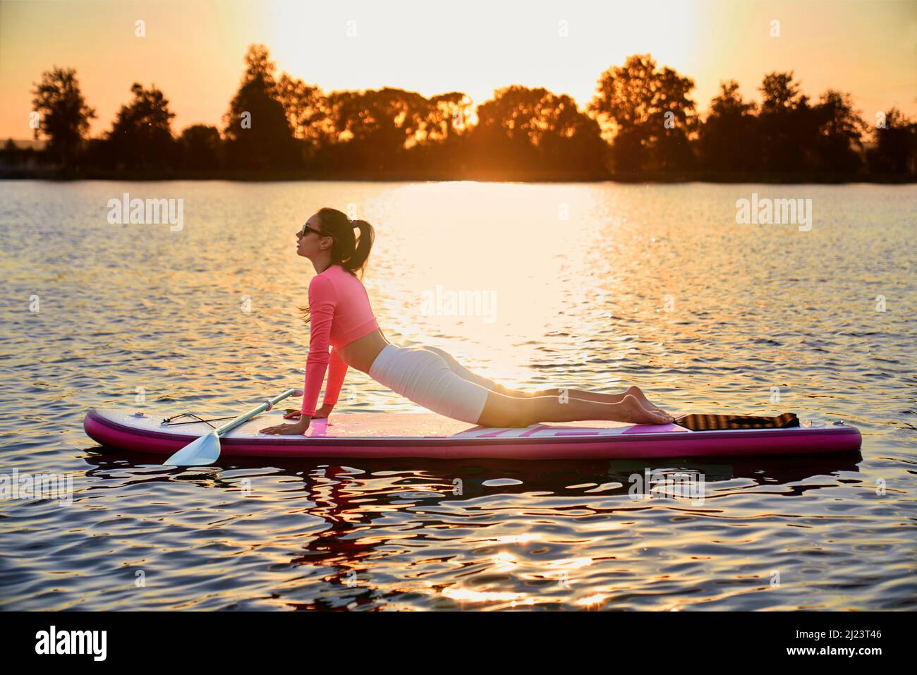 Fitness junge Frau in Sportswear Stretching Body auf Paddle Board. Kaukasische Brünette macht flexible Übungen während des Sonnenuntergangs über dem See. Stockfoto