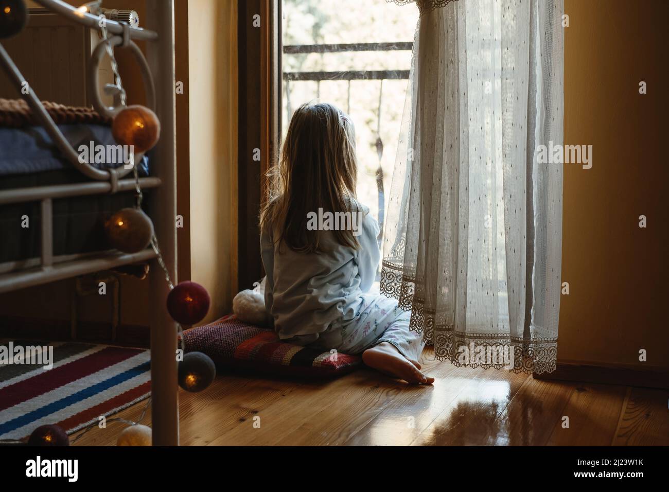 Kind sitzt auf dem Boden und spielt Ameise nach Hause. Verträumte, sonnige Stimmung. Mädchen, das von hinten durch das Fenster schaut. Niedliches Kind wartet darauf, spazieren zu gehen Stockfoto