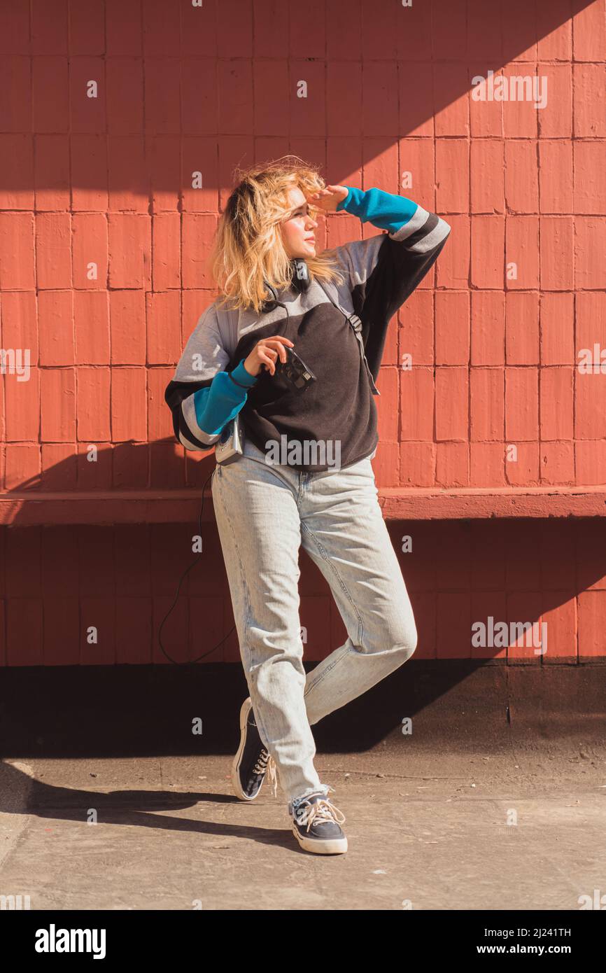 Schöne 90s-inspirierte junge Erwachsene Frau in der Sonne, die ihre Augen schützt, während sie eine altmodische Digitalkamera hält. Hochwertige Fotos Stockfoto