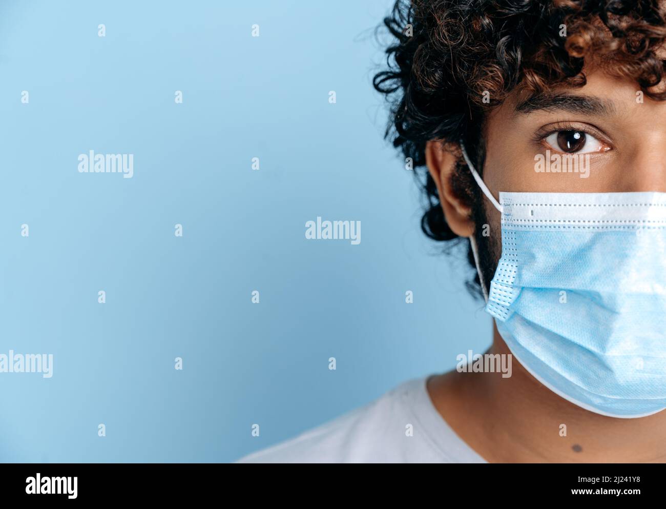 Infektionsprävention. Das halbe Gesicht eines jungen indischen oder arabischen Erwachsenen mit lockiger Kerls, mit einer schützenden medizinischen Maske auf seinem Gesicht, der auf die Kamera blickt, während er auf einem isolierten blauen Hintergrund steht Stockfoto