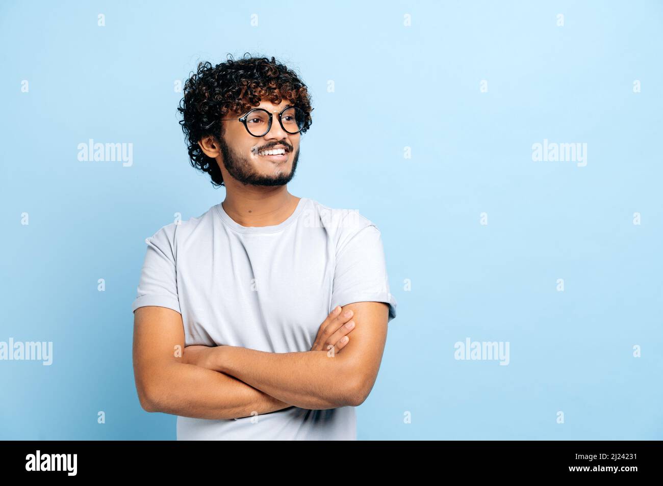 Attraktiver, positiver indischer oder arabischer lockiger Kerl mit Brille, in einem einfachen T-Shirt, Student oder Freiberufler, auf einem isolierten blauen Hintergrund stehend, mit gekreuzten Armen, sieht zur Seite, lächelt Stockfoto