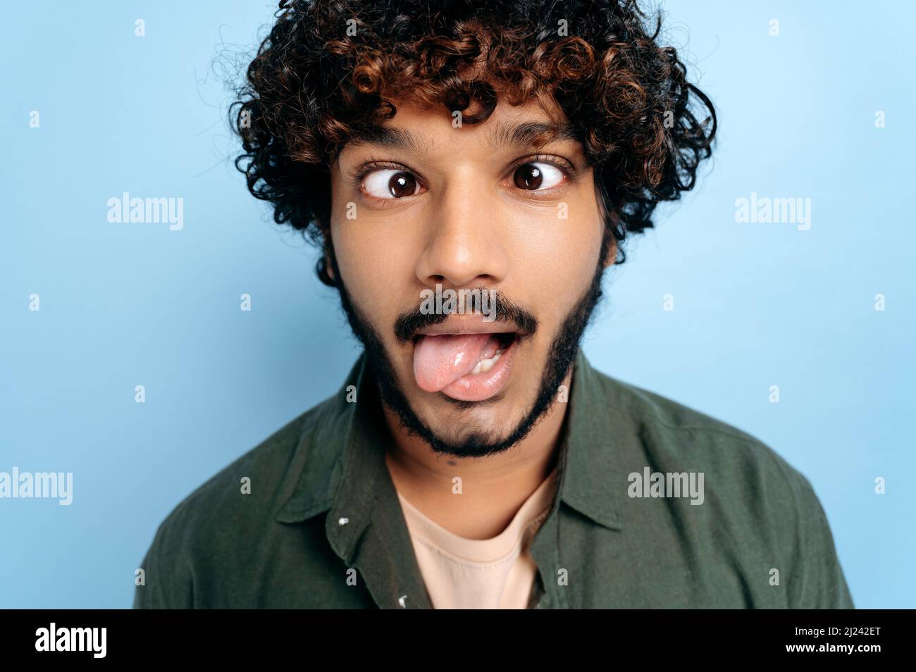 Nahaufnahme des indischen oder arabischen lockigen lustigen Typen, der ein lustiges Gesicht machte, die Zunge heraus, seine Augen schielte, auf isoliertem blauem Hintergrund stehend. Grimassen, die sich herumtäuschen Stockfoto