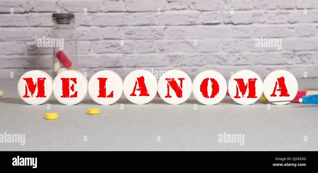Auf blauem Hintergrund ein Stethoskop, eine Spritze und Pillen sowie ein Holzblock mit dem Wort MELANOMA. Medizinisches Konzept Stockfoto