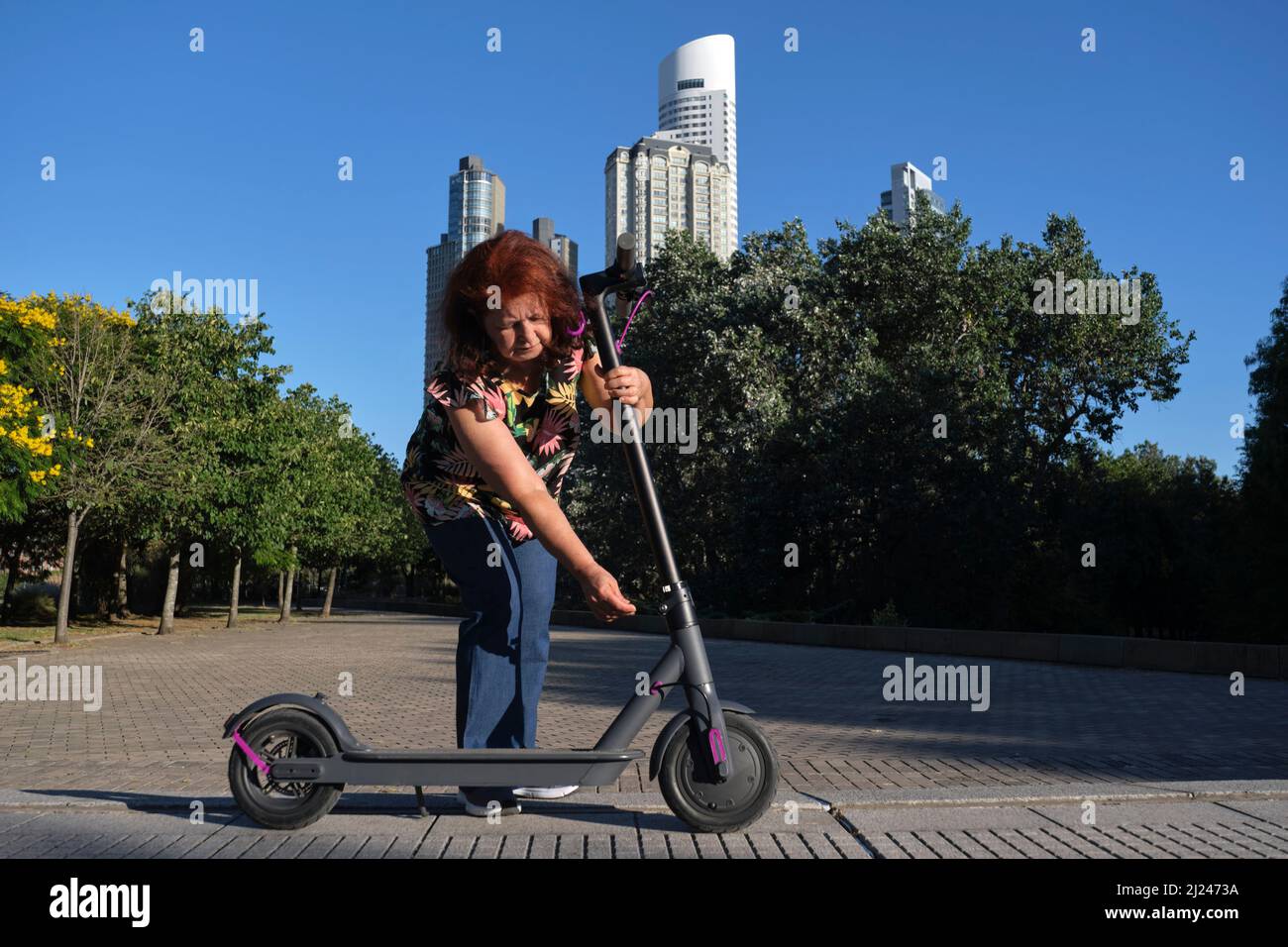 Hispanische reife Frau entfaltet ihren elektrischen Kick Scooter in einem Stadtpark an einem sonnigen Morgen. Konzepte der elektrischen Energie, grüne Mobilität, Öko-Freund Stockfoto