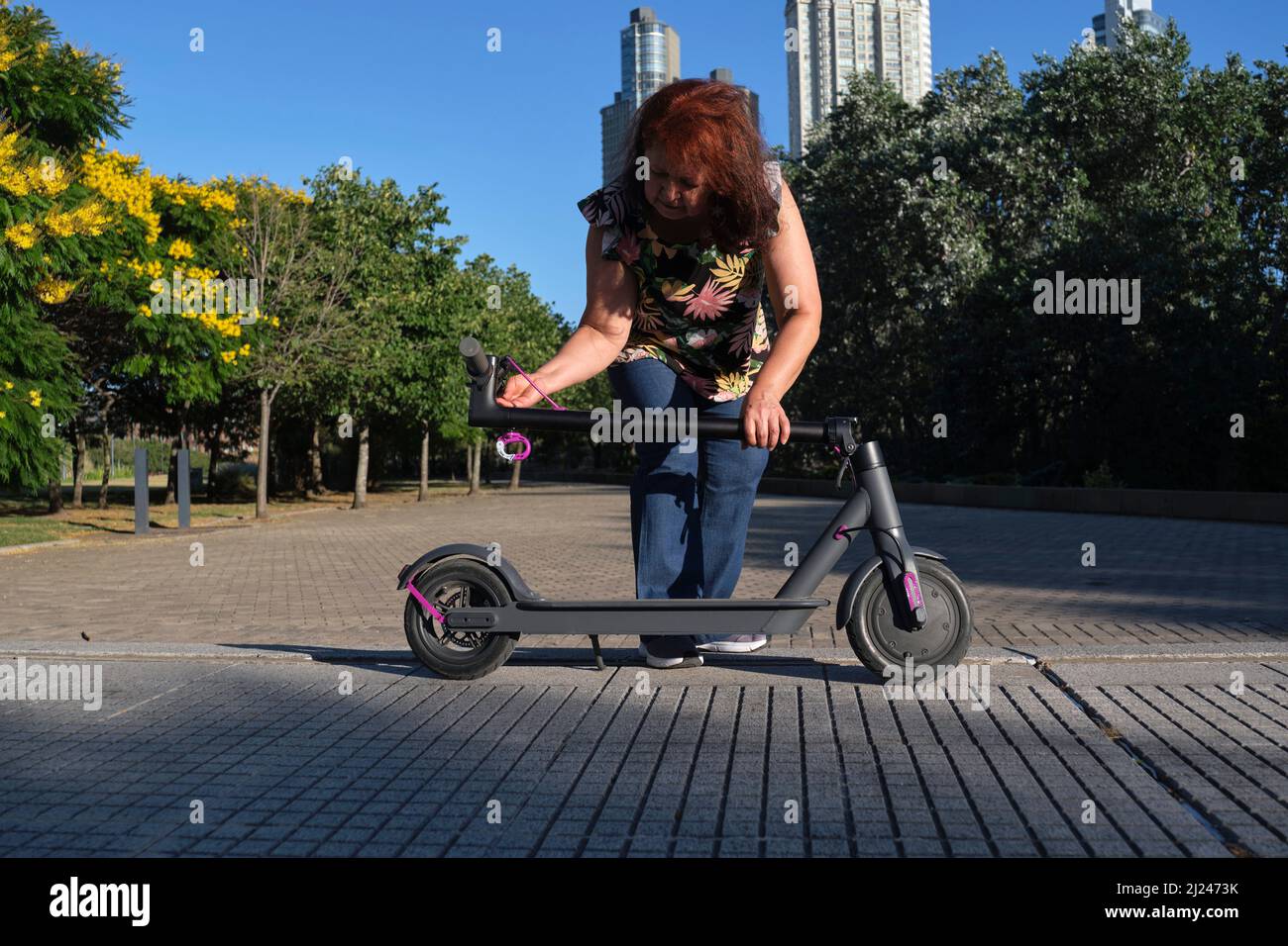 Hispanische reife Frau entfaltet ihren elektrischen Kick Scooter in einem Stadtpark an einem sonnigen Morgen. Konzepte der elektrischen Energie, grüne Mobilität, Öko-Freund Stockfoto