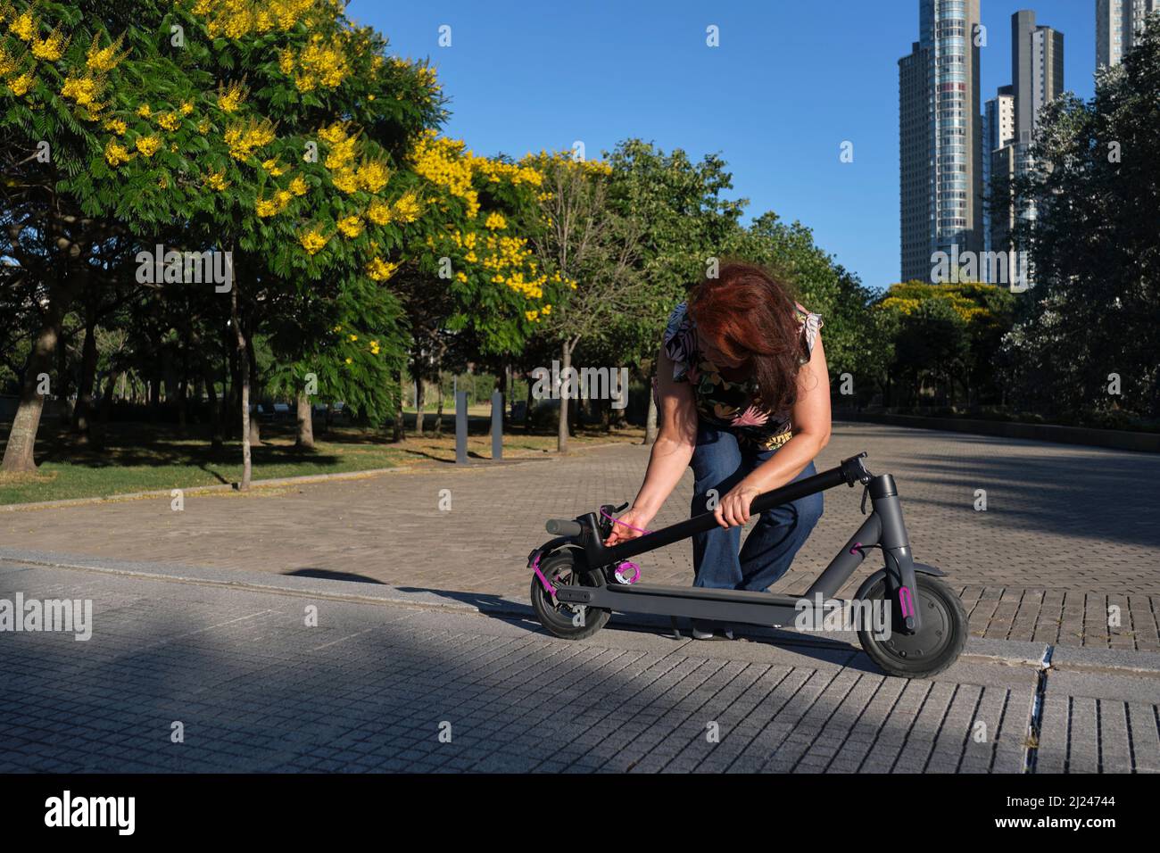 Eine reife Frau aus Latein entfaltet ihren Elektroroller an einem sonnigen Morgen in einem Stadtpark. Konzepte der elektrischen Energie, grüne Mobilität, umweltfreundlich Stockfoto