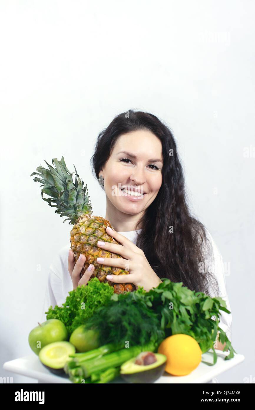 Eine lächelnde Frau mit Gemüse und Obst. Ananas, Äpfel und Sellerie. Das Konzept von Frühling und Vitaminen. Entgiften von Lebensmitteln. Vorderansicht. Stockfoto