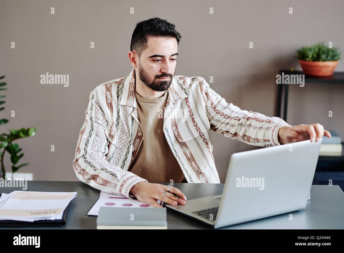 Seriöser ceo in legerer Kleidung, der Laptop öffnet und den Bildschirm anschaut, während er sich für ein digitales Meeting am Schreibtisch im Büro im Freiraum vorbereitete Stockfoto
