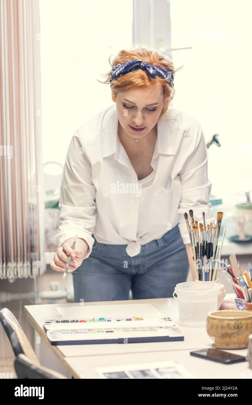 Eine junge, aktive Künstlerin mit roten Haaren, in einem weißen Hemd und Jeans mit einem Verband auf dem Kopf, malt am Tisch Stockfoto