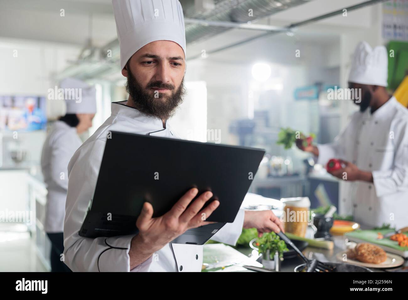 Arbeiter in der Lebensmittelindustrie mit Laptop, nach Gourmet-Gericht Rezept auf dem Bildschirm rühren Zutaten in der Pfanne. Chefkoch mit Computer, der die Garnierung für das Hauptgericht vorbereitet, während er in der Restaurantküche ist. Stockfoto