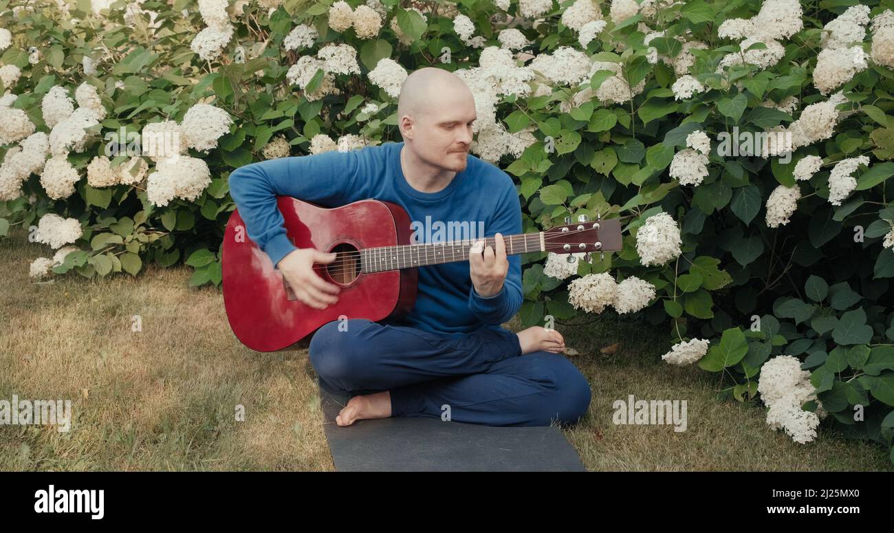 Der kaukasische Mann sitzt in einem Park zwischen Blumen und spielt rote Akustikgitarre Stockfoto