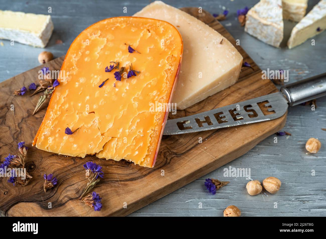 Käsehintergrund. Geschnittenes Stück Käse, Messer mit Aufschrift 'Cheese', Mutter auf Holzschneidebrett auf Tisch. Stockfoto