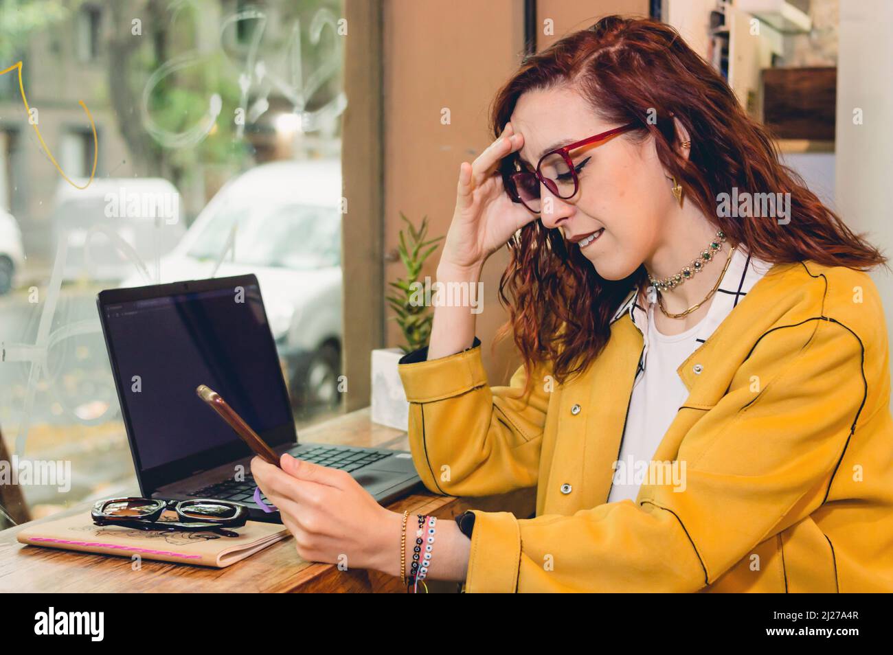 Rothaarige junge Frau mit Brille und gelbem Blazer freiberuflich mit ihrem Laptop in einem Café arbeiten und empört auf das Telefon schauen. Stockfoto