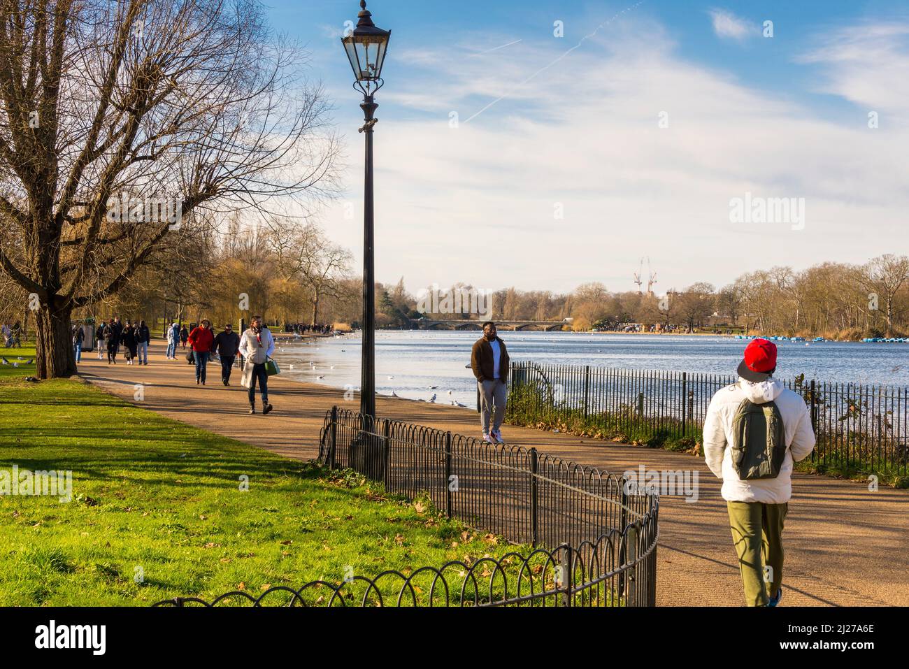 Menschen, die an einem altmodischen Laternenpfosten auf dem Fußweg am Serpentine Lake im Hyde Park, London, an einem hellen sonnigen Wintertag spazieren gehen. Stockfoto