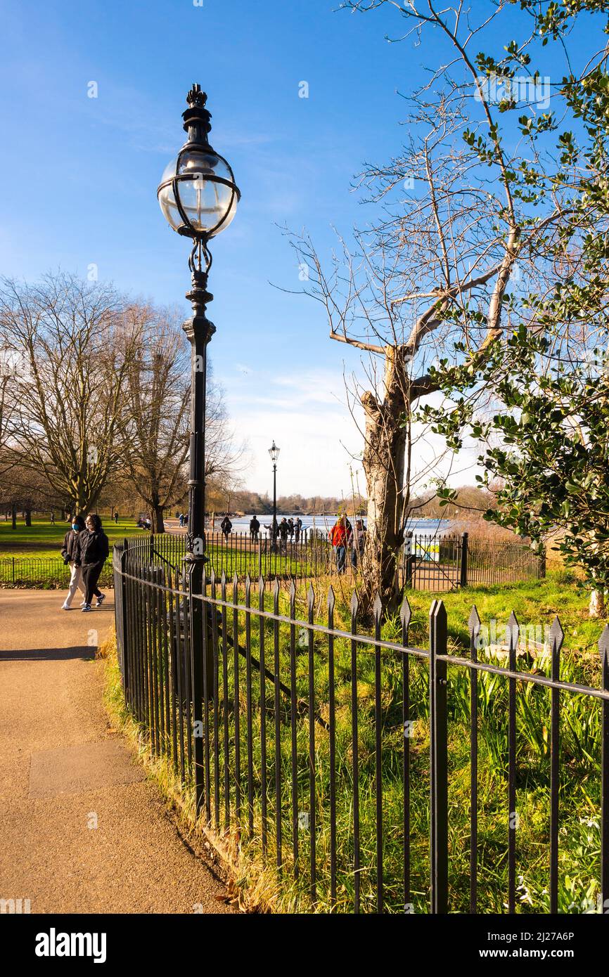 Menschen, die an einem altmodischen Laternenpfosten auf dem Fußweg am Serpentine Lake im Hyde Park, London, an einem hellen sonnigen Wintertag spazieren gehen. Stockfoto