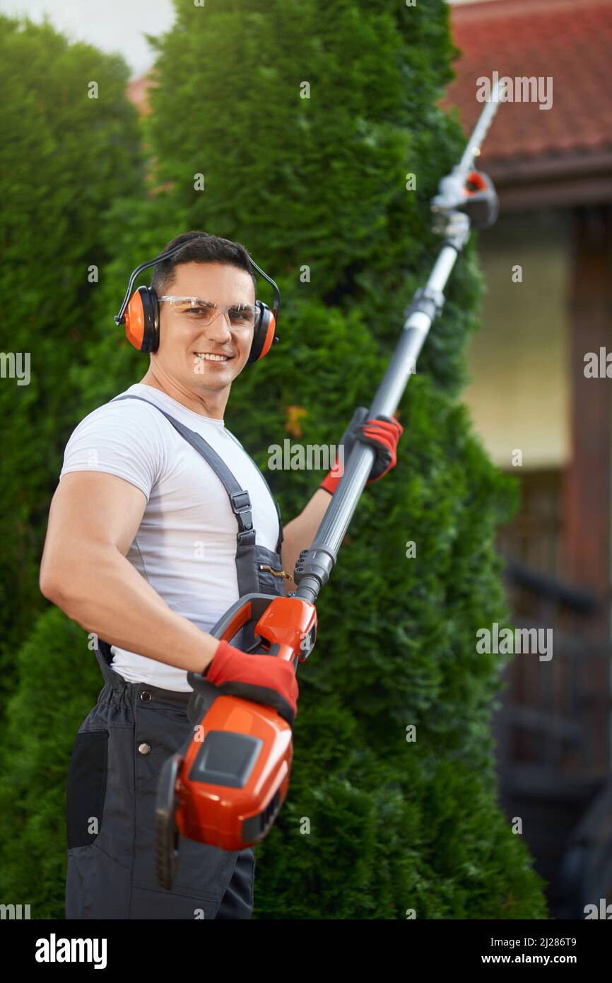 Lächelnder Gärtner in Uniform, Schutzbrille, Kopfhörer und Handschuhe halten die Benzin-Heckenschere, während er auf dem Hinterhof steht. Kaukasischer Mann mit modernen Gartengeräten im Freien. Stockfoto