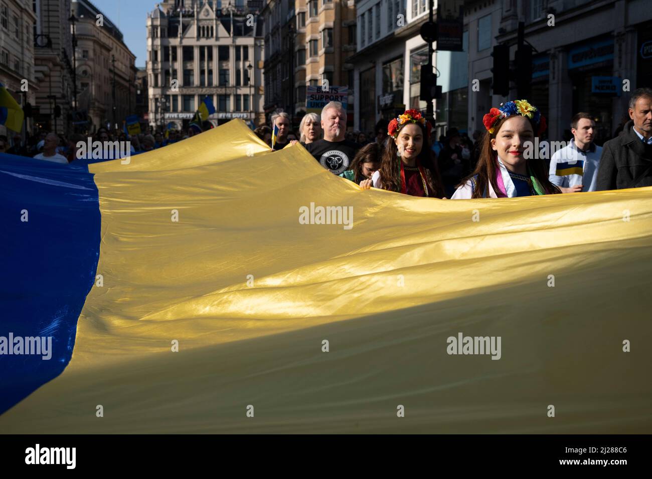 Junge Frauen mit traditionellen Blumen im Haar halten eine riesige ukrainische Flagge, während sich Tausende von Menschen am 26.. März 2022 in London, Großbritannien, in der Hauptstadt zu einem friedensmarsch ‘London steht mit der Ukraine’ versammeln. Der Konflikt in der Ukraine befindet sich nun in seinem zweiten Monat, und ihr Präsident hat die Menschen auf der ganzen Welt dazu aufgerufen, sich gegen die russische Invasion und den anhaltenden Krieg in der Region zu vereinen. Die ukrainische Flagge in Gelb und Blau war überall in dieser bunten Show der Einheit zu sehen. Stockfoto