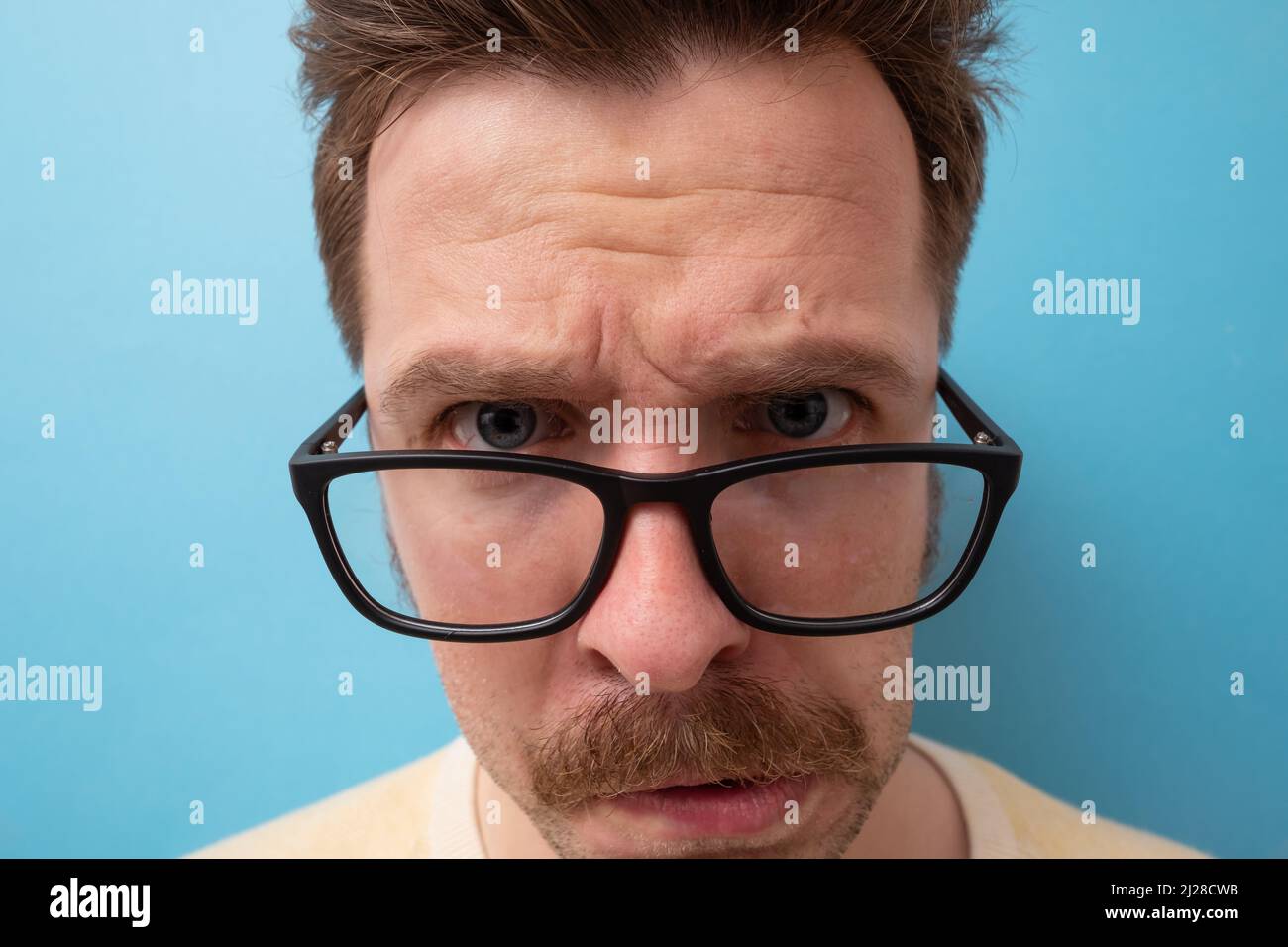 Junger Mann mit witzigem Schnurrbart in großer Brille, der Zweifel hat und mit verwirrem Gesichtsausdruck auf isoliertem blauem Hintergrund Stockfoto