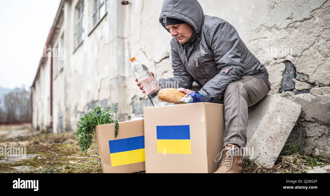 Ein älterer Mann in warmen Kleidern sitzt am Rande eines entgleisten Gebäudes und nimmt Brot und Wasser aus dem humanitären Hilfspaket für die Ukraine. Stockfoto