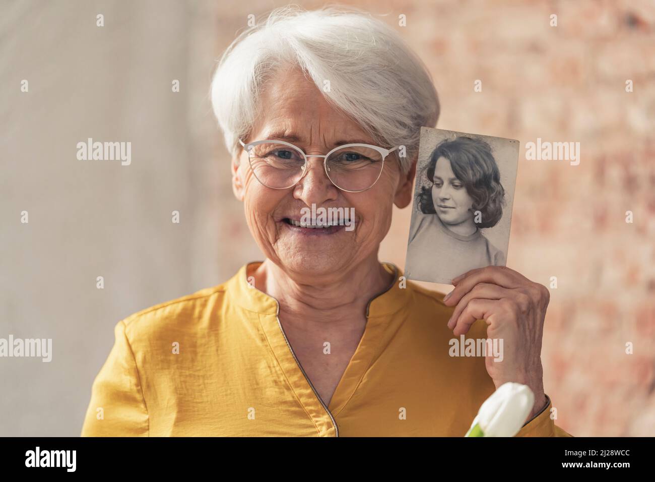 Lächelnde ältere Dame mit kurzen grauen Haaren vergleicht ihr aktuelles Gesicht mit einem Foto ihres jüngeren selbst. Hochwertige Fotos Stockfoto
