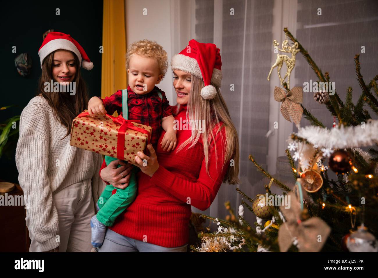 Eine Mutter und eine große Schwester schenken einem zweijährigen Kind zu Weihnachten ein Geschenk. Stockfoto