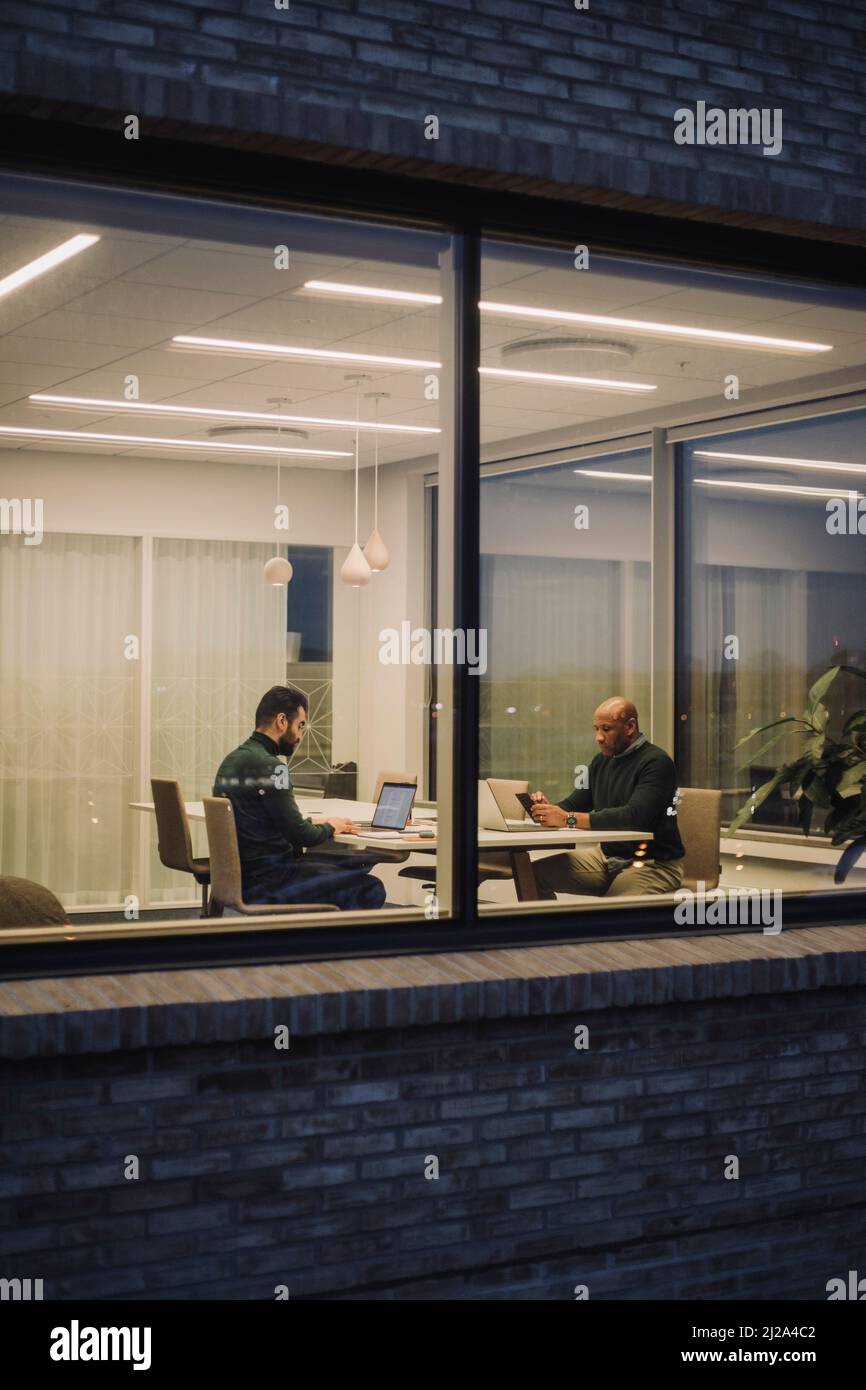 Ein Geschäftsmann mit einem Kollegen, der spät im Büro arbeitet und nachts durch das Fenster gesehen wird Stockfoto