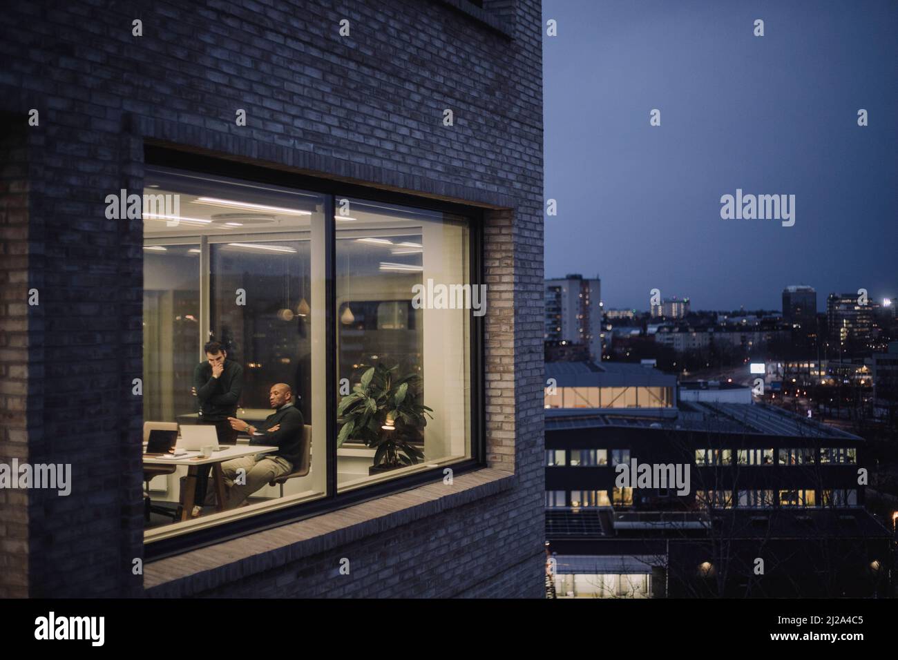 Reifer Geschäftsmann, der mit einem männlichen Kollegen über einen Laptop spricht, während er nachts im Büro Überstunden arbeitet Stockfoto
