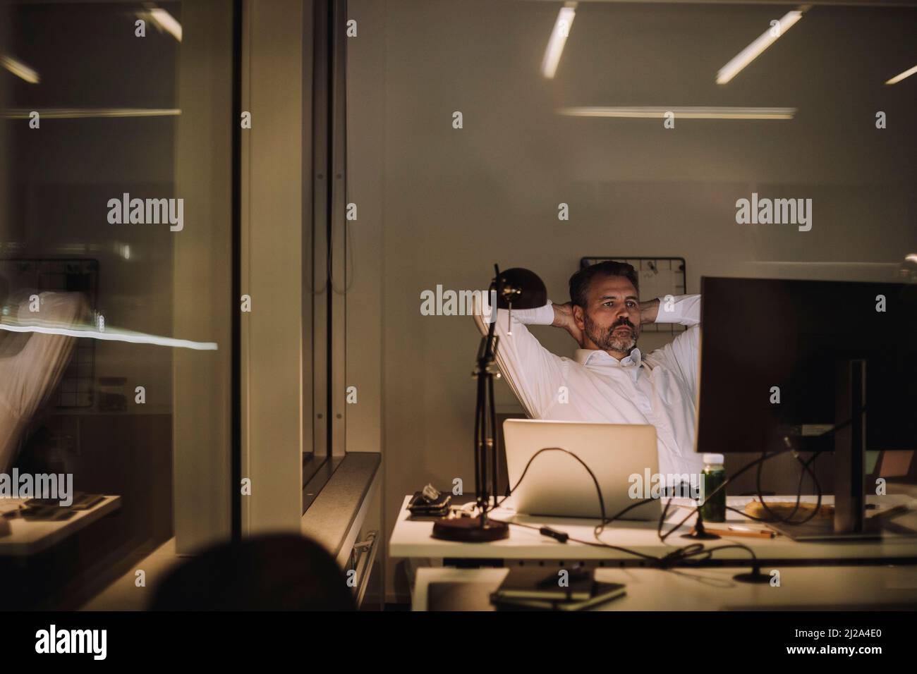 Reifer Geschäftsmann mit Händen hinter dem Kopf, der nachts im Büro arbeitet Stockfoto