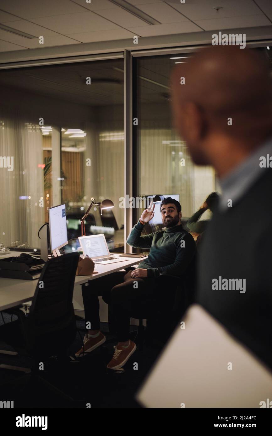 Geschäftsmann, der mit einem männlichen Kollegen spricht, während er nachts im Büro Überstunden arbeitet Stockfoto